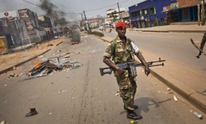 A heavily armed military policeman patrols next to burning barricades set up by angry supporters of opposition leader Kizza Besigye, near to his party headquarters, in Kampala, Uganda, Friday, Feb. 19, 2016. Police in Uganda arrested opposition leader Kizza Besigye at his party's headquarters Friday after heavily armed police surrounded the building and fired tear gas and stun grenades at his supporters who took to the streets. (AP Photo/Ben Curtis)