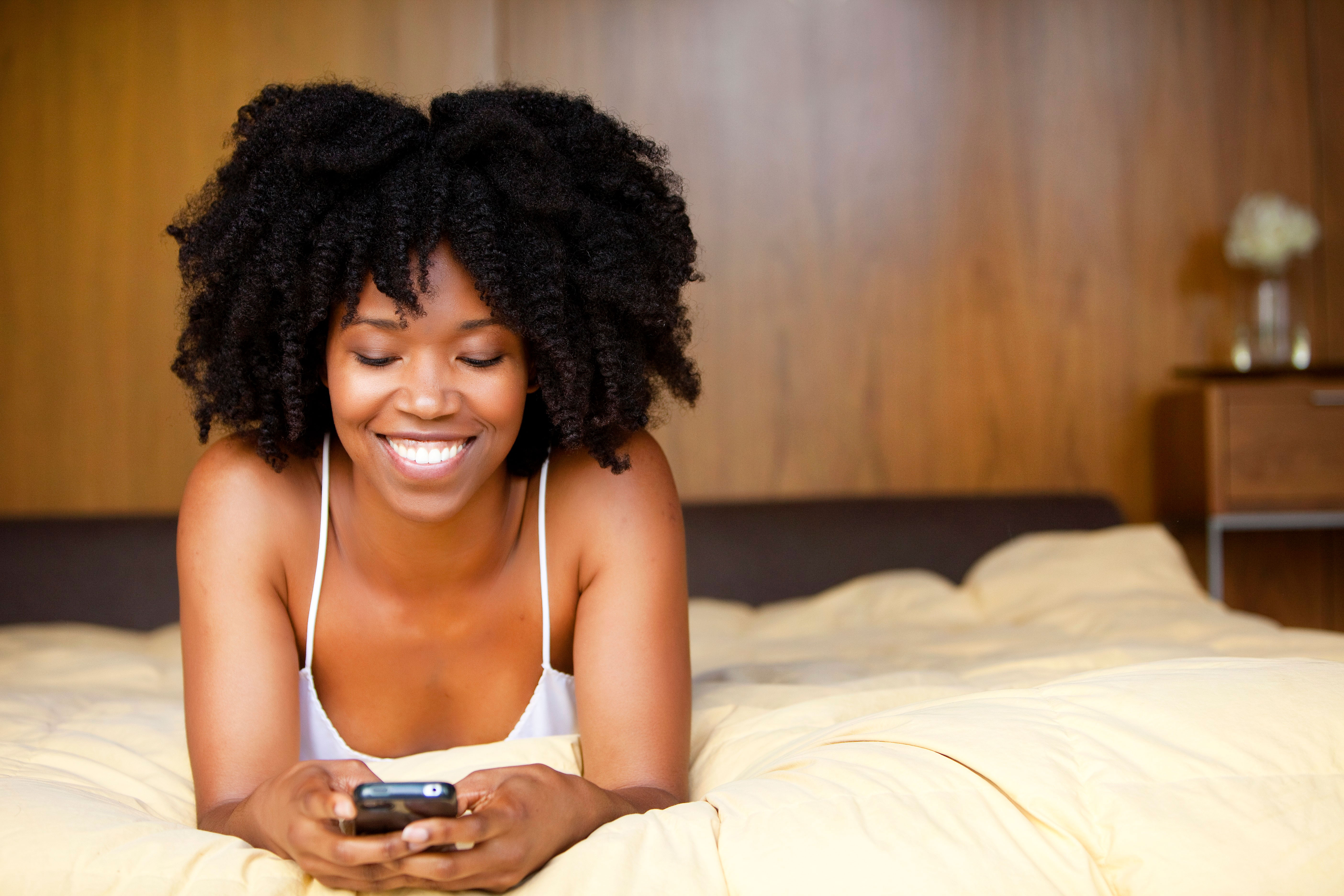 A black woman lying in bed using a cell phone. --- Image by © Michael Poehlman/Corbis
